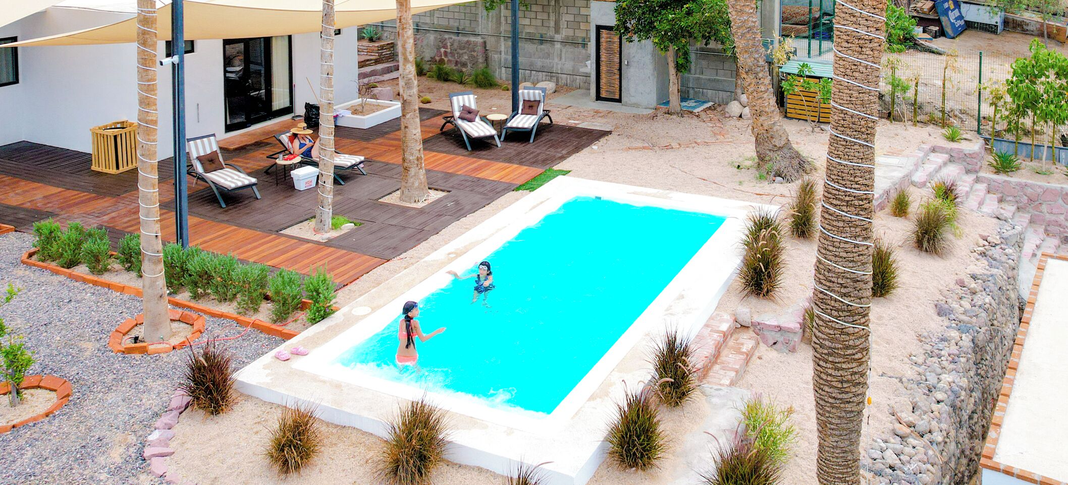 Une piscine rectangulaire où se trouvent deux personnes, entourée d’une terrasse aménagée avec des chaises longues et une petite table.