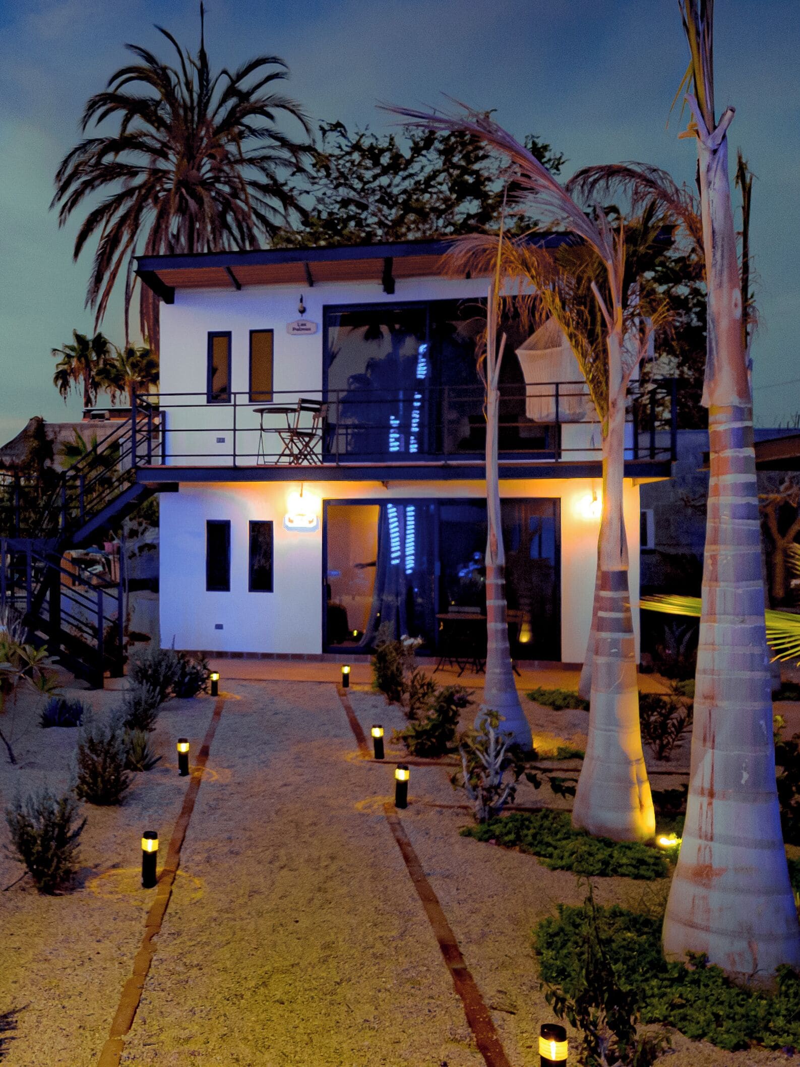 A modern two-story building with a balcony, surrounded by palm trees and a well-lit pathway.