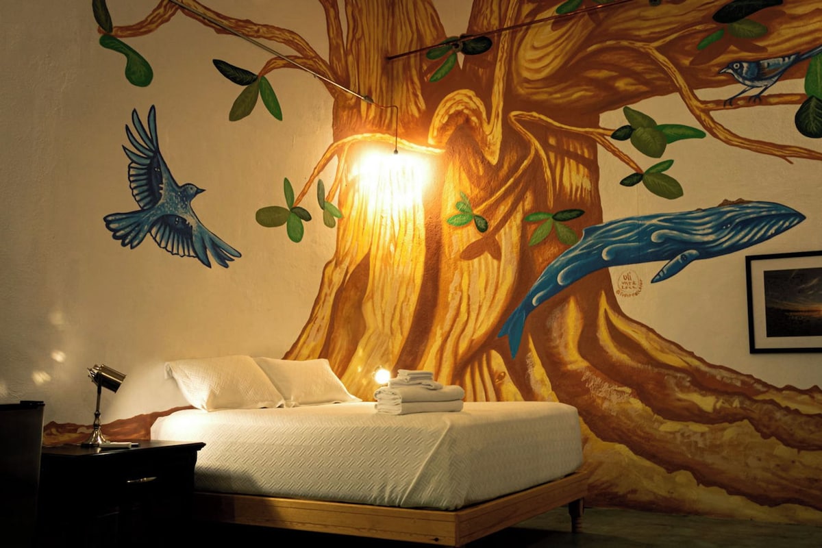 Une chambre avec un grand décor mural représentant un arbre, un lit et un bureau avec une lampe.