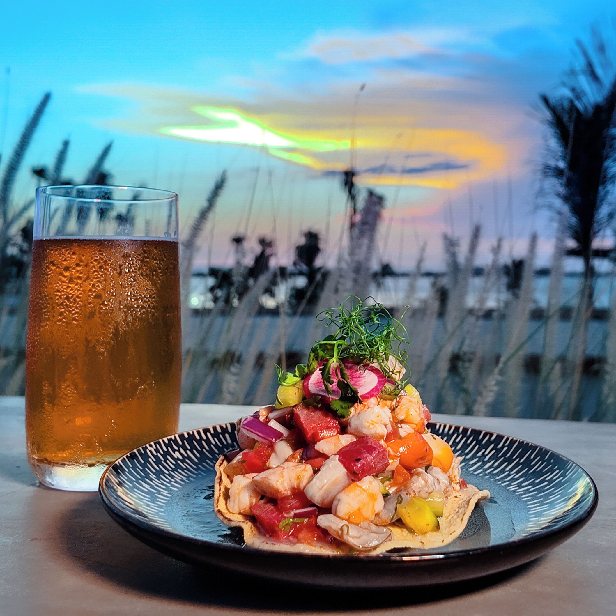 A meal with shrimp and vegetables on a plate, a glass of iced tea, and a view of the beach with trees in the background.