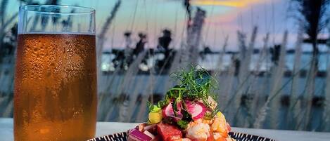 Un plato con camarones y verduras, un vaso de té helado y vista a la playa con árboles al fondo.