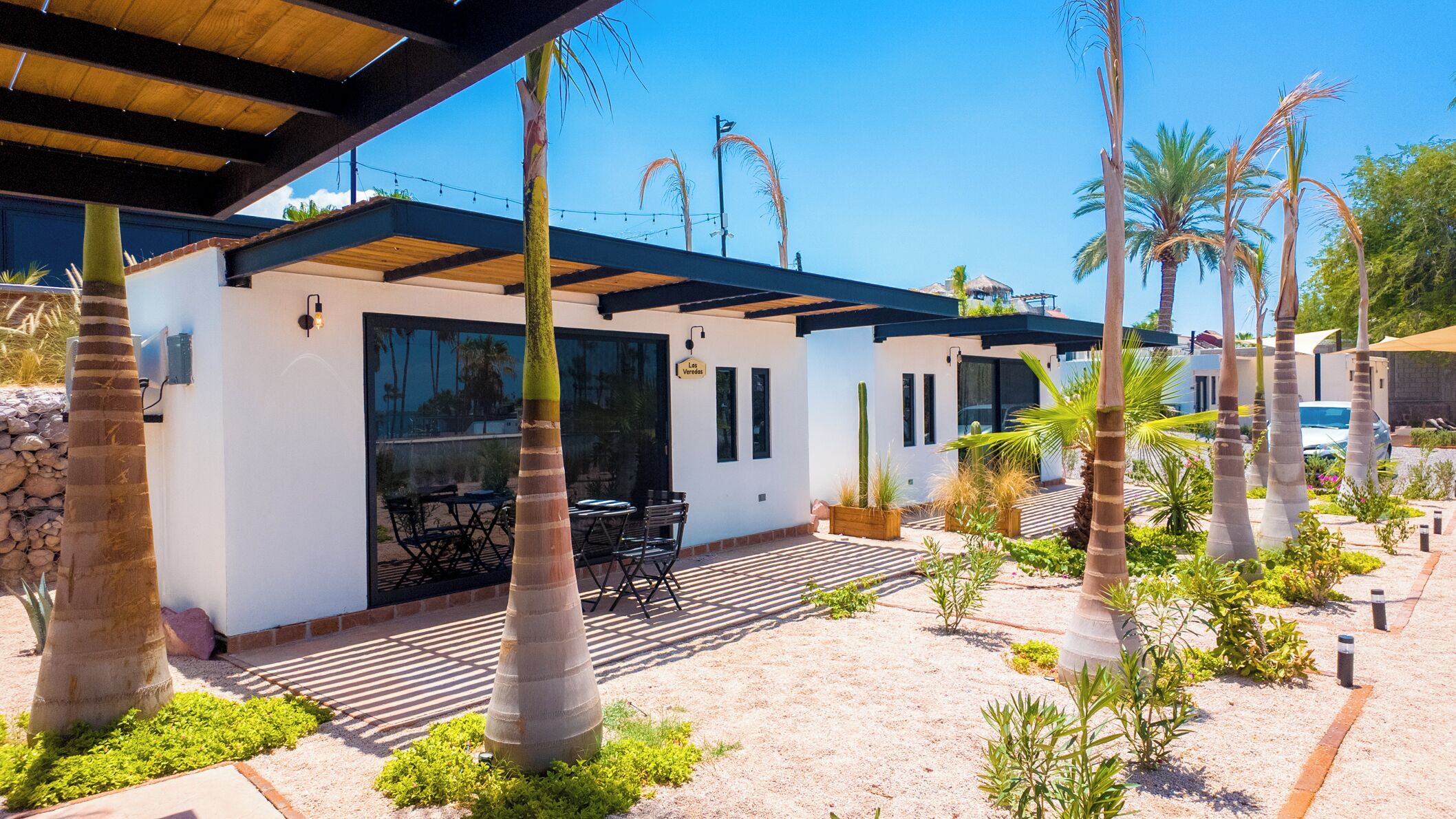 Un patio moderno al aire libre con zona de comedor, palmeras y un edificio blanco con un letrero que dice "La Vereda".