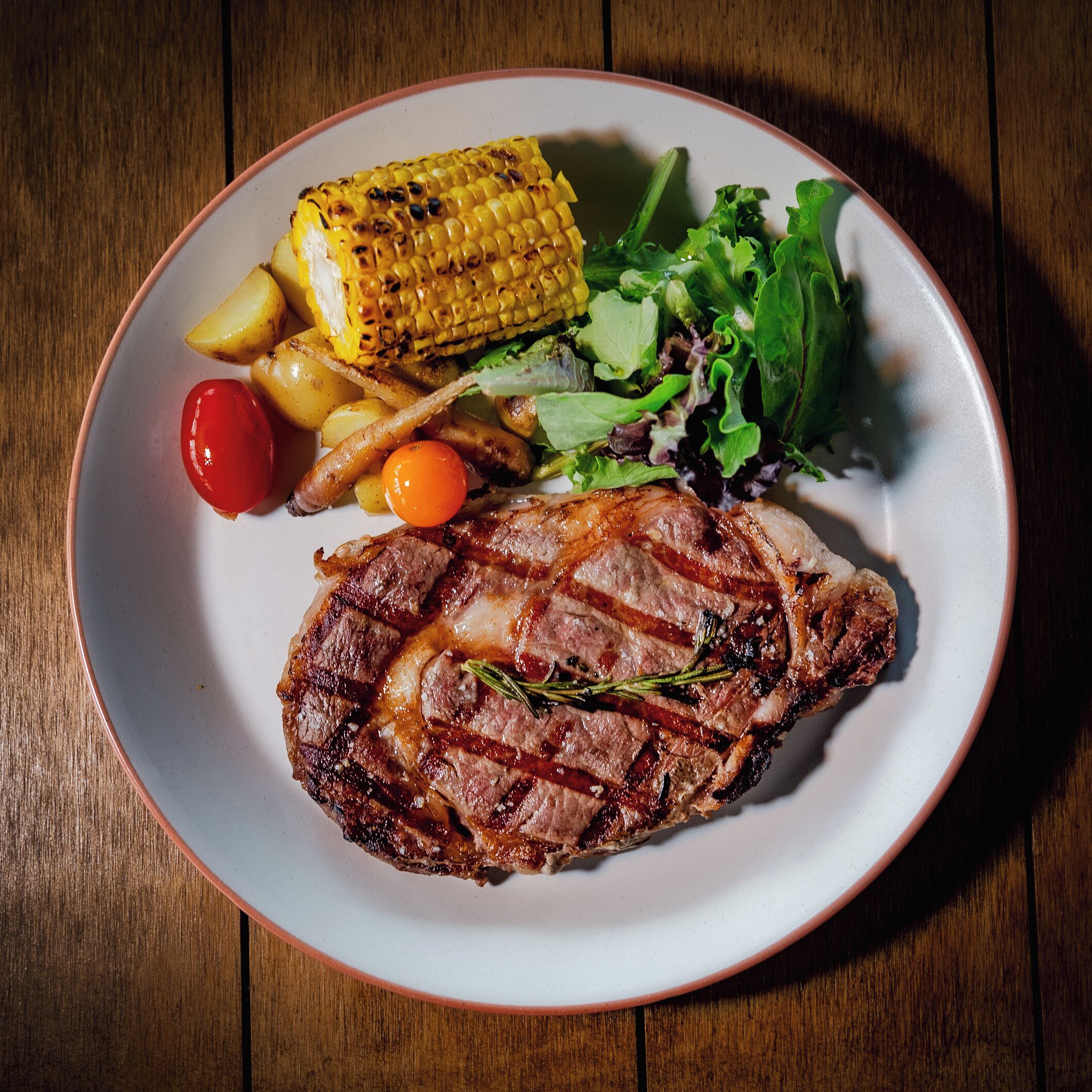 Un plato con bistec a la parrilla, mazorca de maíz, patatas asadas, tomates cherry y una ensalada.