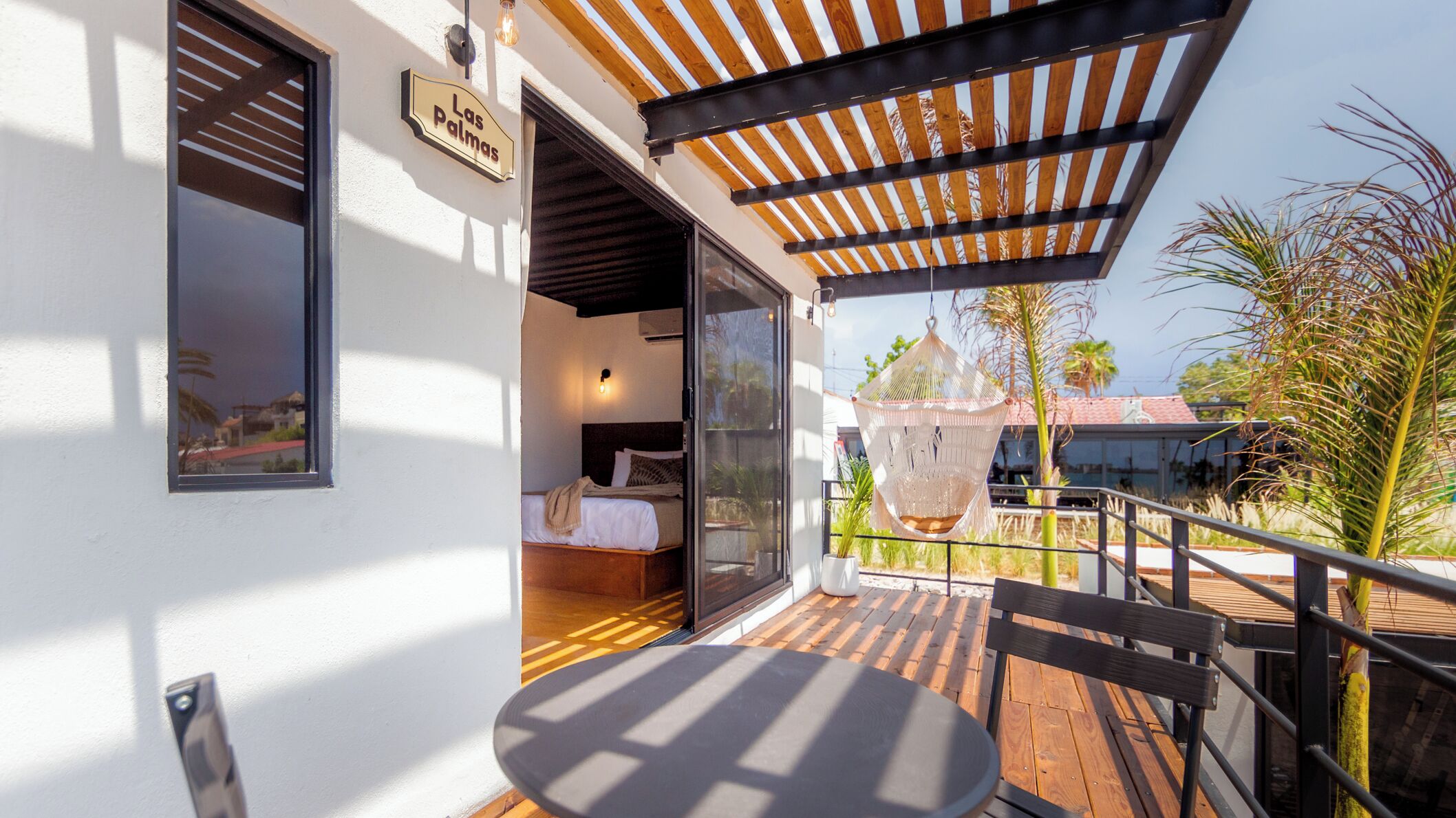 Un balcón con mesa y sillas de madera, una silla colgante y una cama visible a través de una puerta abierta.