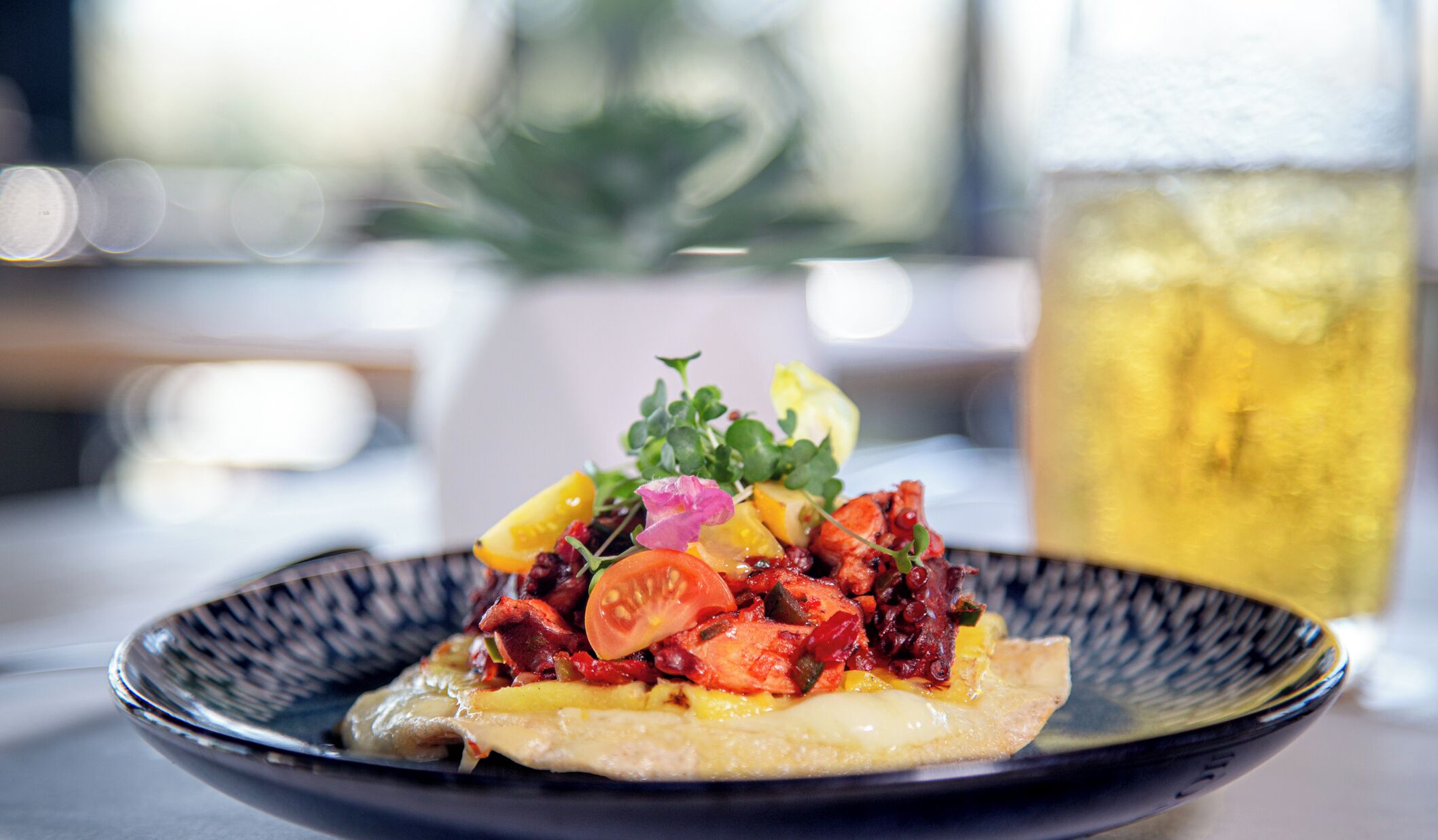 A dish with various vegetables and a drink in the background.