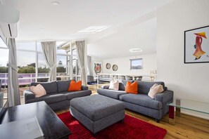 Una sala de estar moderna con sofás grises, cojines rojo y naranja, una mesa de centro de vidrio y una alfombra roja. Hay un ventanal con cortinas, un cuadro de un tigre y vistas a la vegetación exterior.