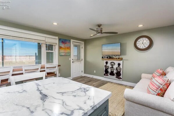 A spacious living room with a marble dining table, a sofa with colorful cushions, a ceiling fan, a wall-mounted TV, and a large clock.