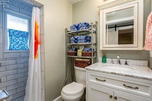 Une salle de bain avec des toilettes blanches, un lavabo en marbre, une étagère avec des paniers et une fenêtre ornée d’un rideau bleu et orange.