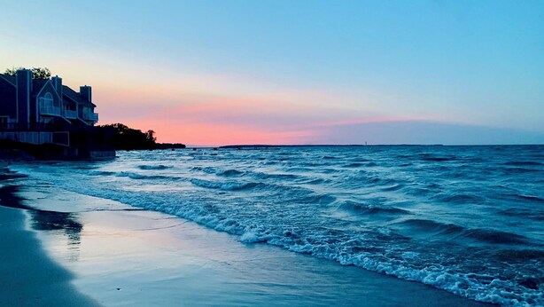 Una playa con olas al atardecer, una casa a la izquierda y un faro en la distancia.