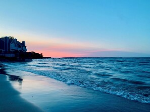 Une plage avec des vagues au coucher du soleil, une maison sur la gauche et un phare au loin.