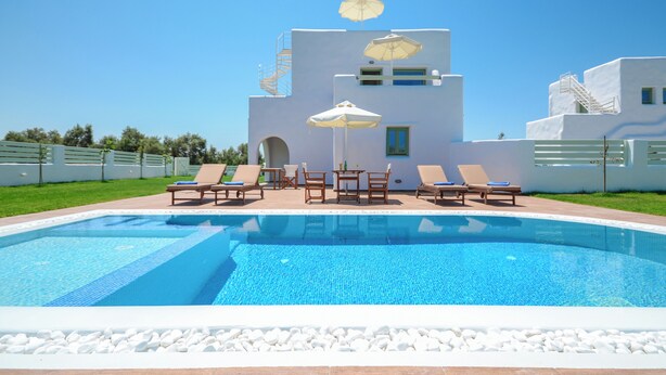 Une piscine avec des chaises longues et des parasols, un bâtiment blanc avec un escalier, et un ciel bleu azur.