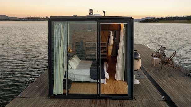 Une chambre à coucher aux murs de verre, avec un lit, des rideaux et une petite table sur une terrasse en bois, le tout donnant sur un plan d’eau.