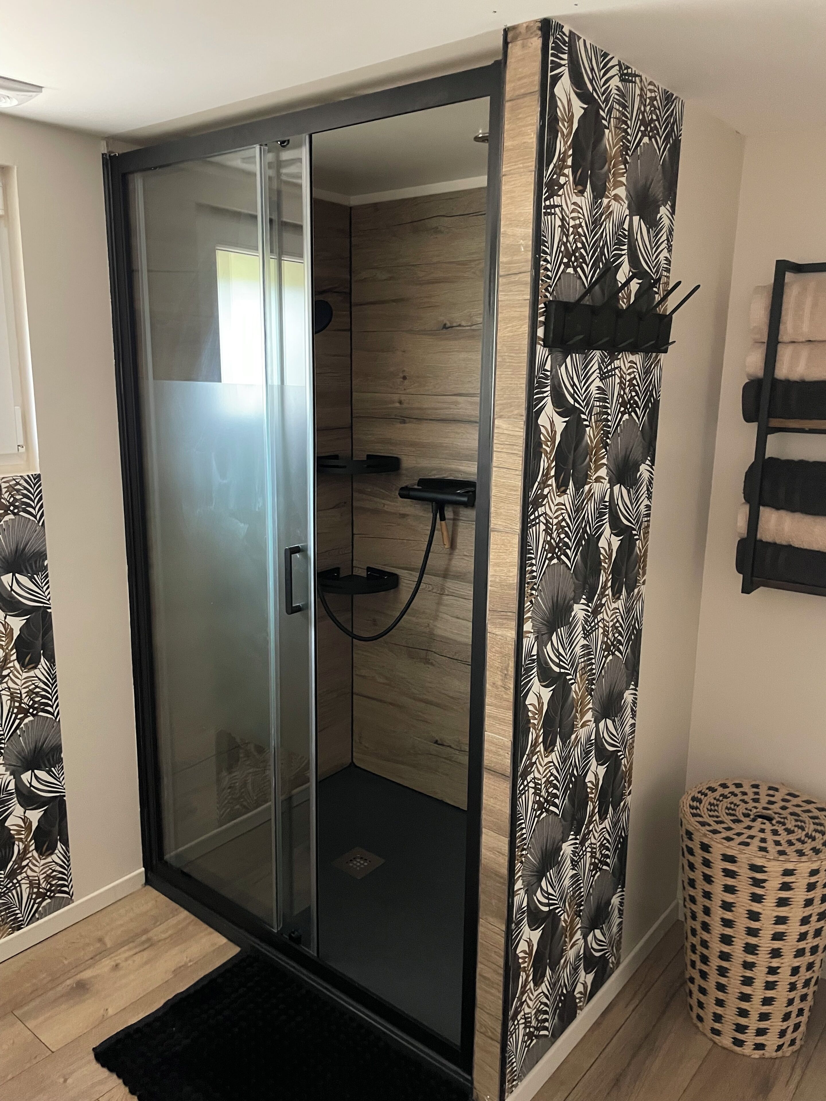 Un espace de douche clos par des parois de verre, avec un intérieur en bois, un tapis noir à l’entrée et un panier tressé posé sur le sol.