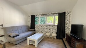 A living room with a grey sofa, a wooden coffee table, two white chairs, a TV on a wooden cabinet, and a window with patterned curtains. - Charming fully-equipped house (La Houssière)