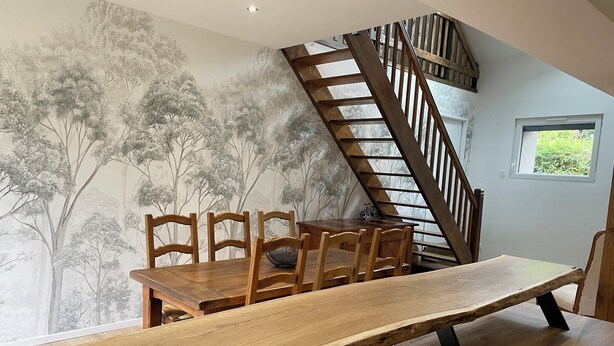 A dining area with a long wooden table and chairs, a staircase, and a mural of trees on the wall. - Charming fully-equipped house (La Houssière)