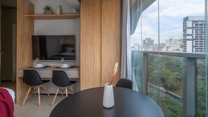 Et moderne spiseområde med et rundt bord, sorte stole og en hvid vase med tørrede planter. Et træskab og et fladskærms-tv ses i baggrunden.