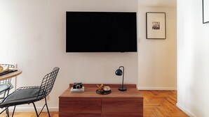 Un soggiorno moderno con una televisione a schermo piatto montata a parete, un tavolino in legno, una sedia nera dal design essenziale, una scrivania con una fotocamera e una lampada, e un quadro appeso al muro.