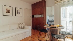 Un ambiente compatto con un divano bianco, un tavolo da pranzo con sedie, un mobiletto in legno e un angolo cottura.