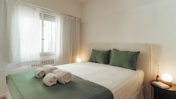 Un letto rifatto con lenzuola verdi, una lampada da comodino e una finestra con tende.