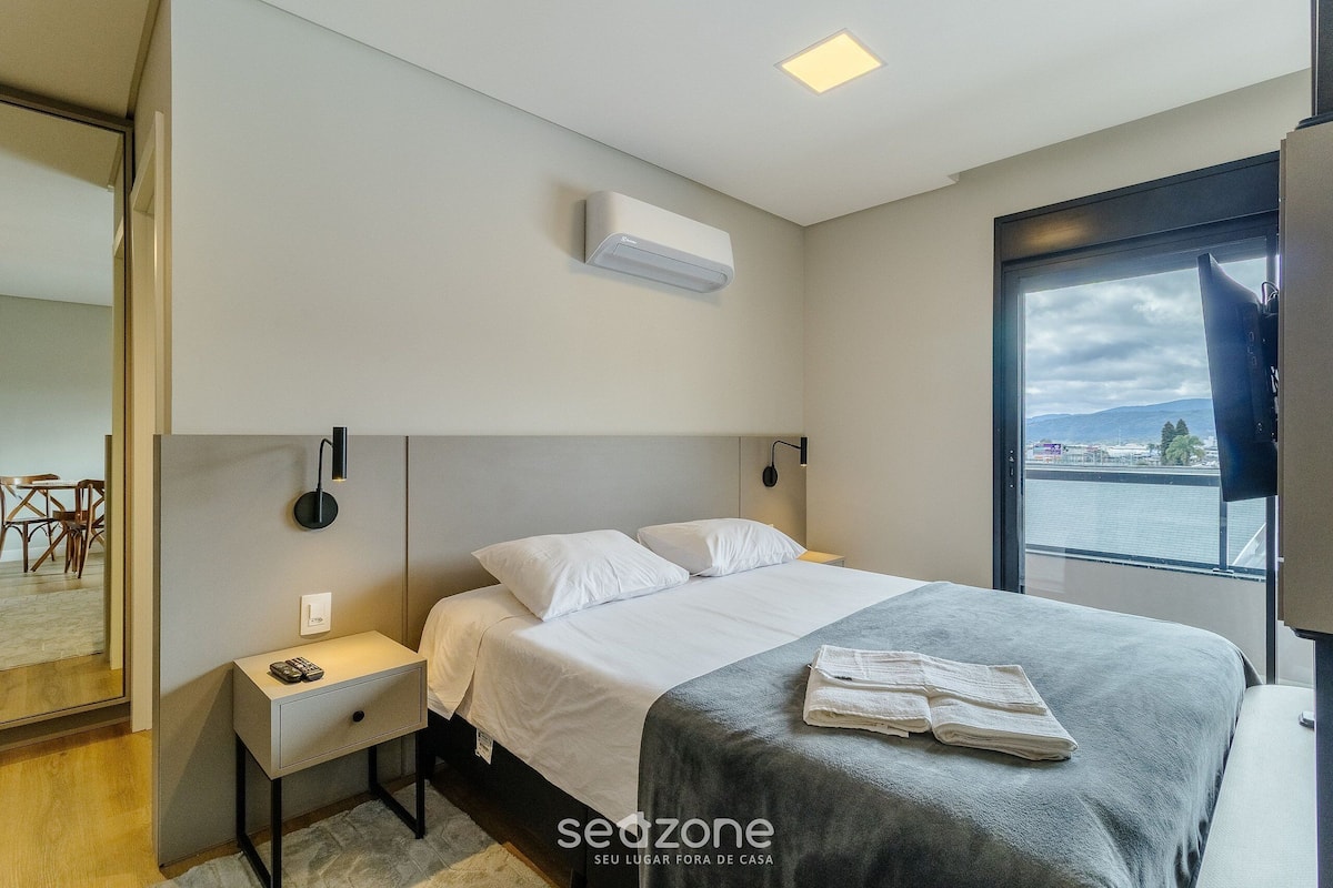 Quarto de hotel moderno com uma cama grande, mesa de cabeceira e vista da cidade através de uma janela ampla.
