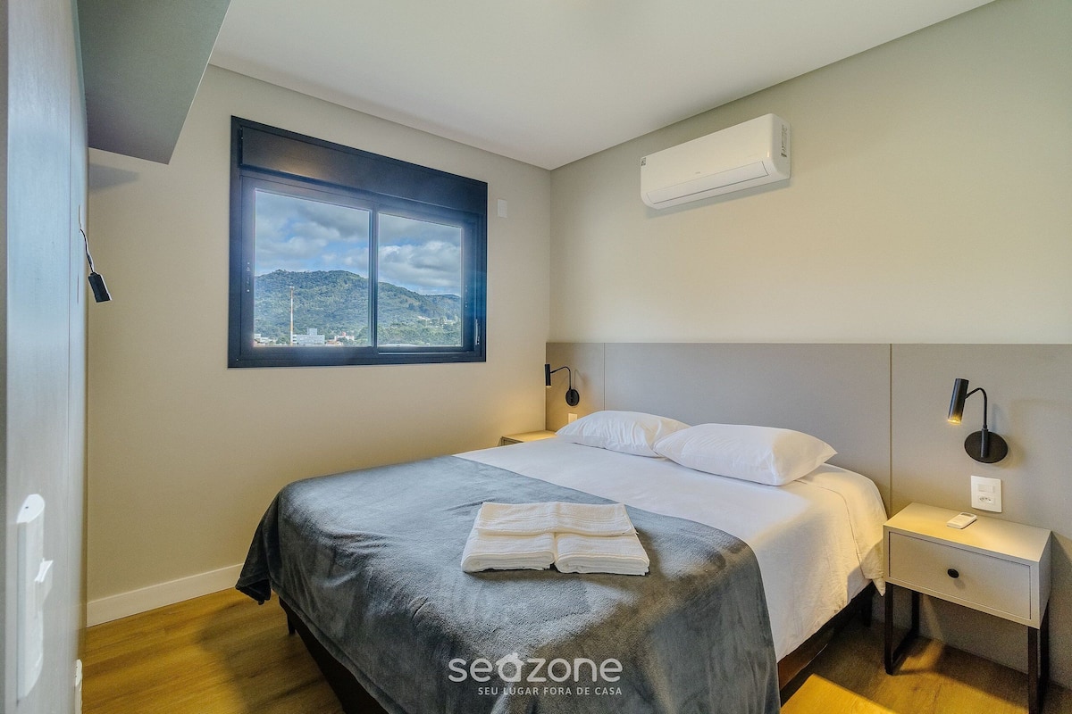 Quarto de hotel moderno com uma cama, mesa de cabeceira, ar condicionado fixado na parede, janela com vista para as montanhas e uma toalha sobre a cama.