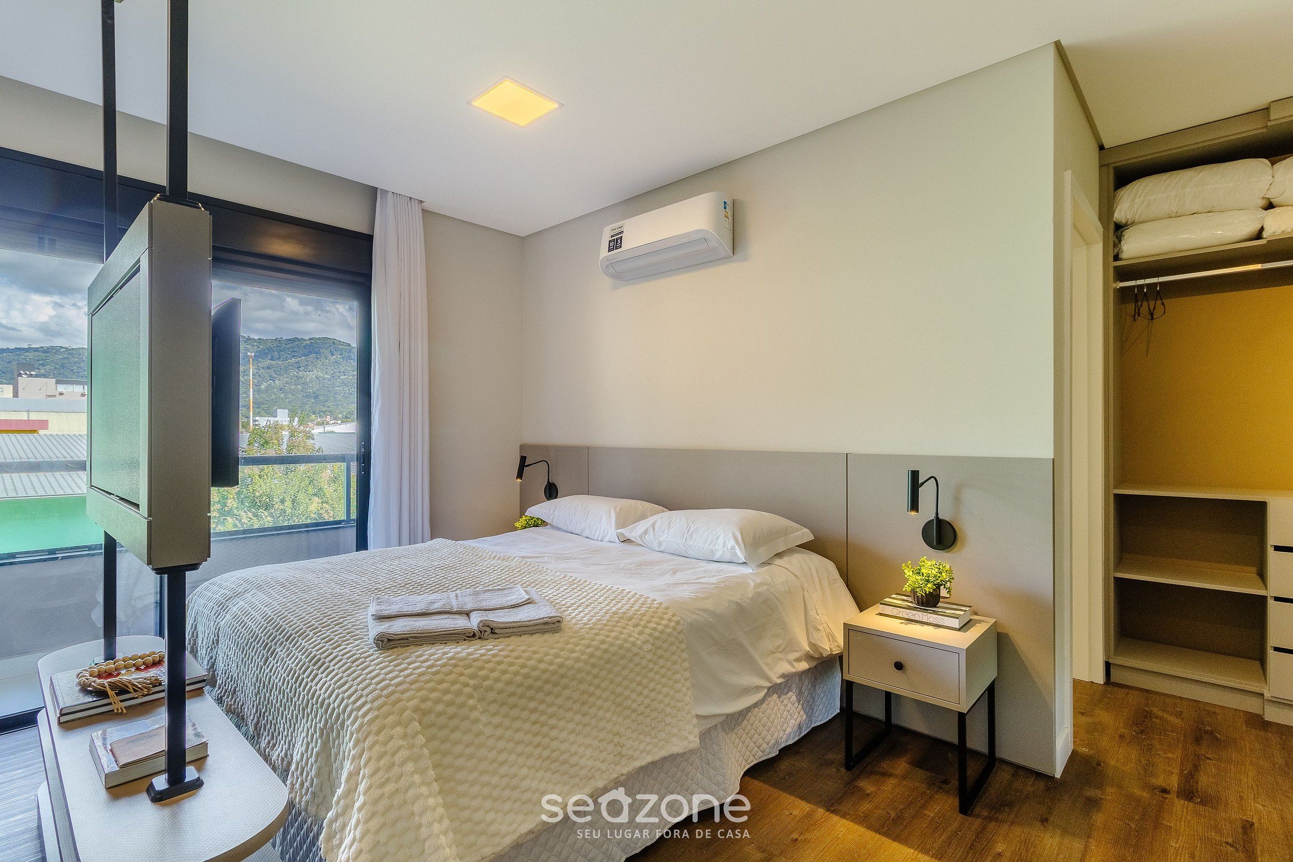 A modern hotel room with a large bed, a TV on a stand, a bedside table with a lamp, and a view of the outdoors.