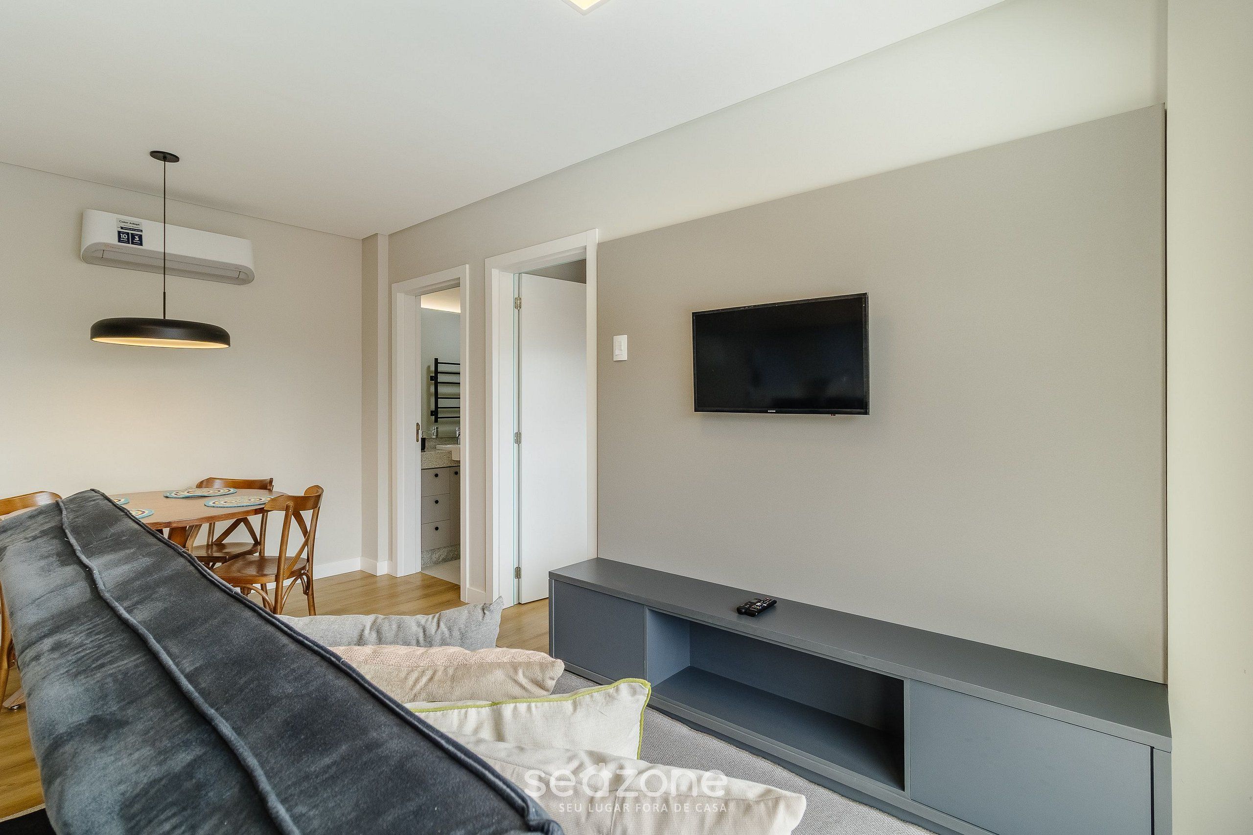 Una sala moderna con un sofá gris, una mesa de comedor con sillas, un televisor de pantalla plana montado en la pared y un banco empotrado debajo del televisor.