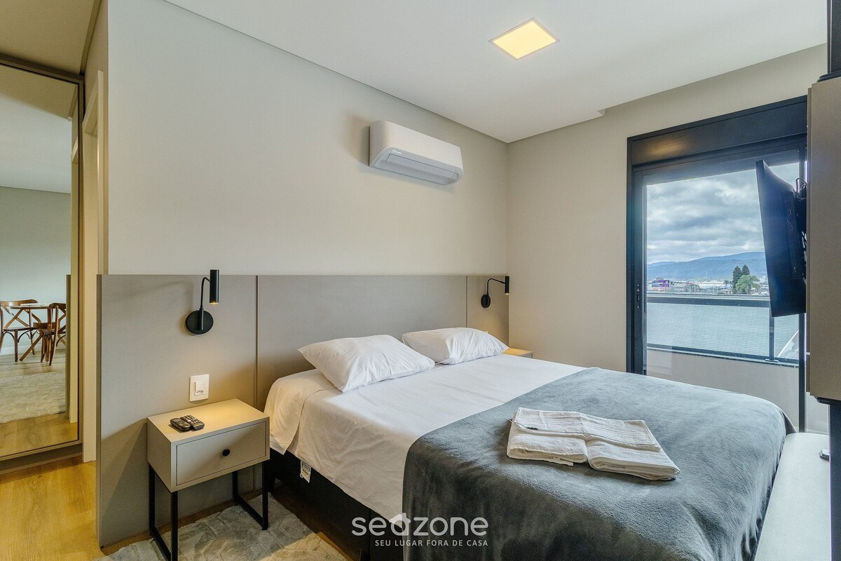 Ett modernt hotellrum med en stor säng, ett sängbord, en liten avlastningsyta och utsikt över staden genom ett stort fönster.