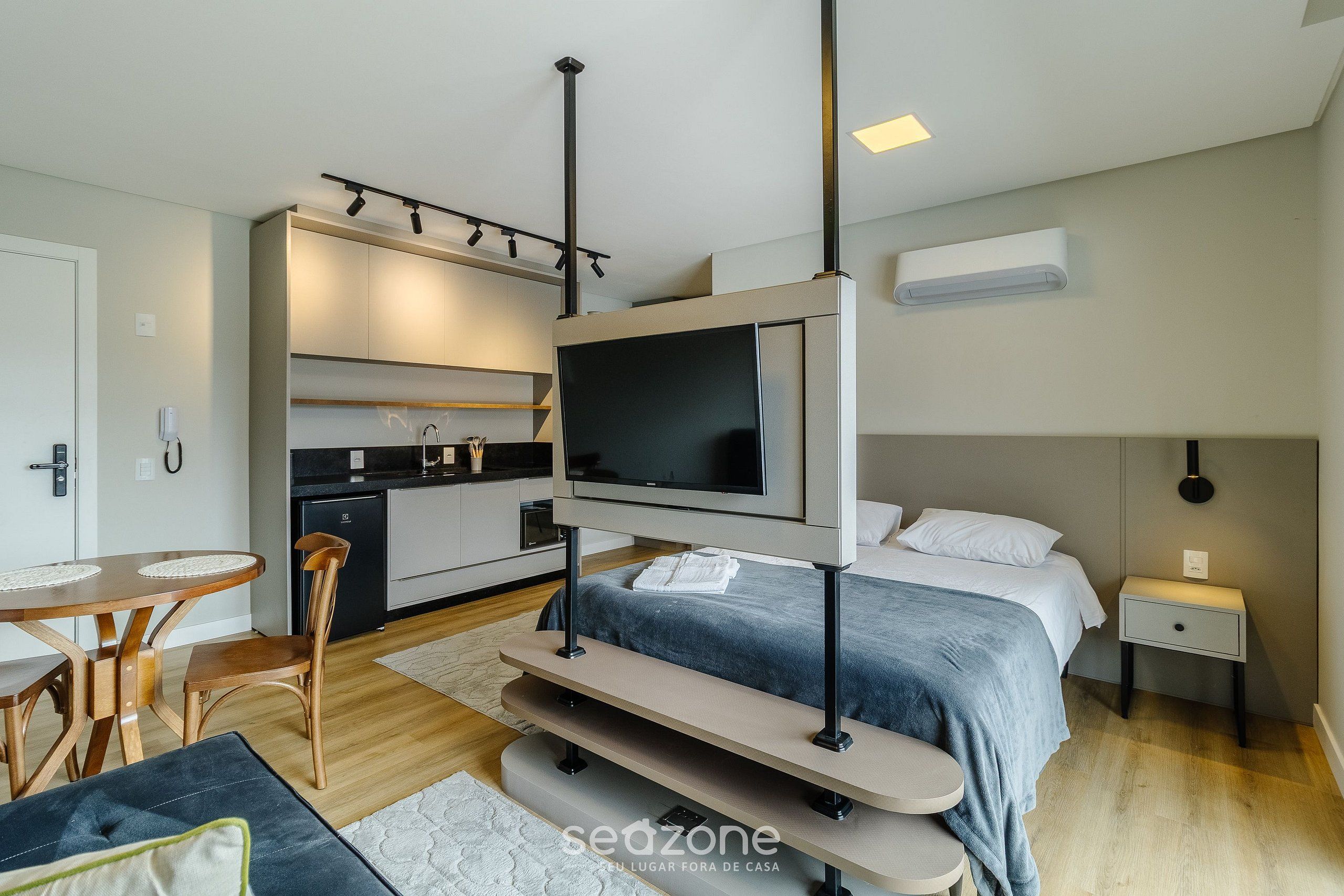 A modern hotel room with a bed, a TV mounted on a stand, a small kitchen area, and a seating area with a table and chairs.