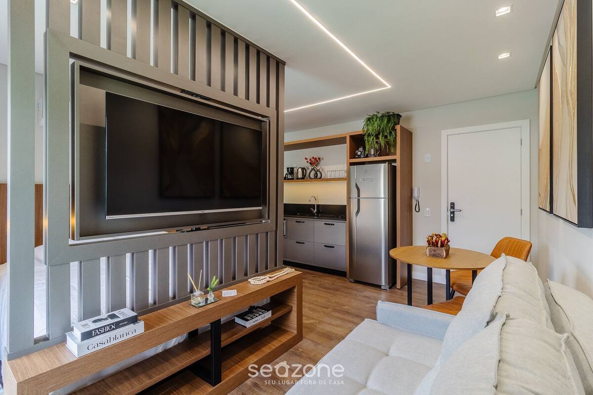 Uma sala de estar moderna com uma televisão de tela plana grande fixada na parede, uma mesa de centro de madeira com livros, um sofá e um espaço de jantar com uma pequena mesa e cadeiras.