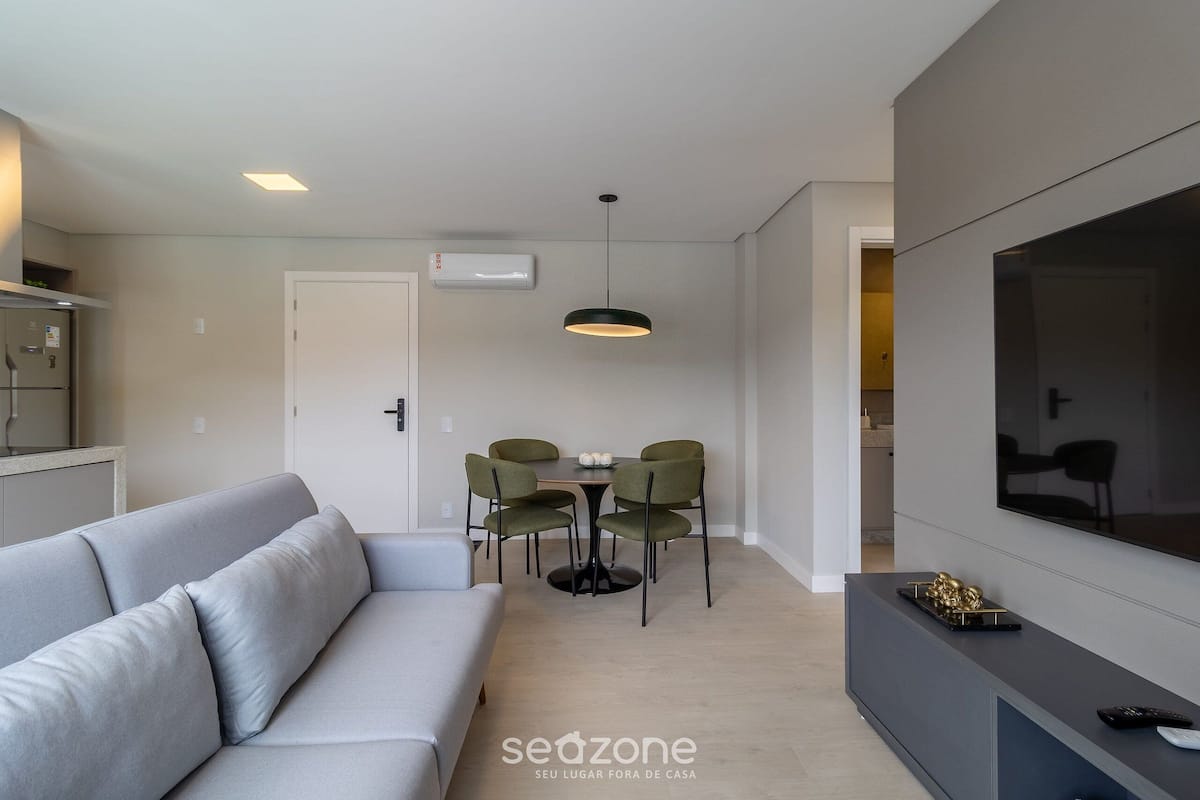 Uma sala de estar moderna com um sofá cinzento, uma mesa de jantar com quatro cadeiras, uma luminária pendente, uma TV sobre um suporte e uma área de cozinha ao fundo.