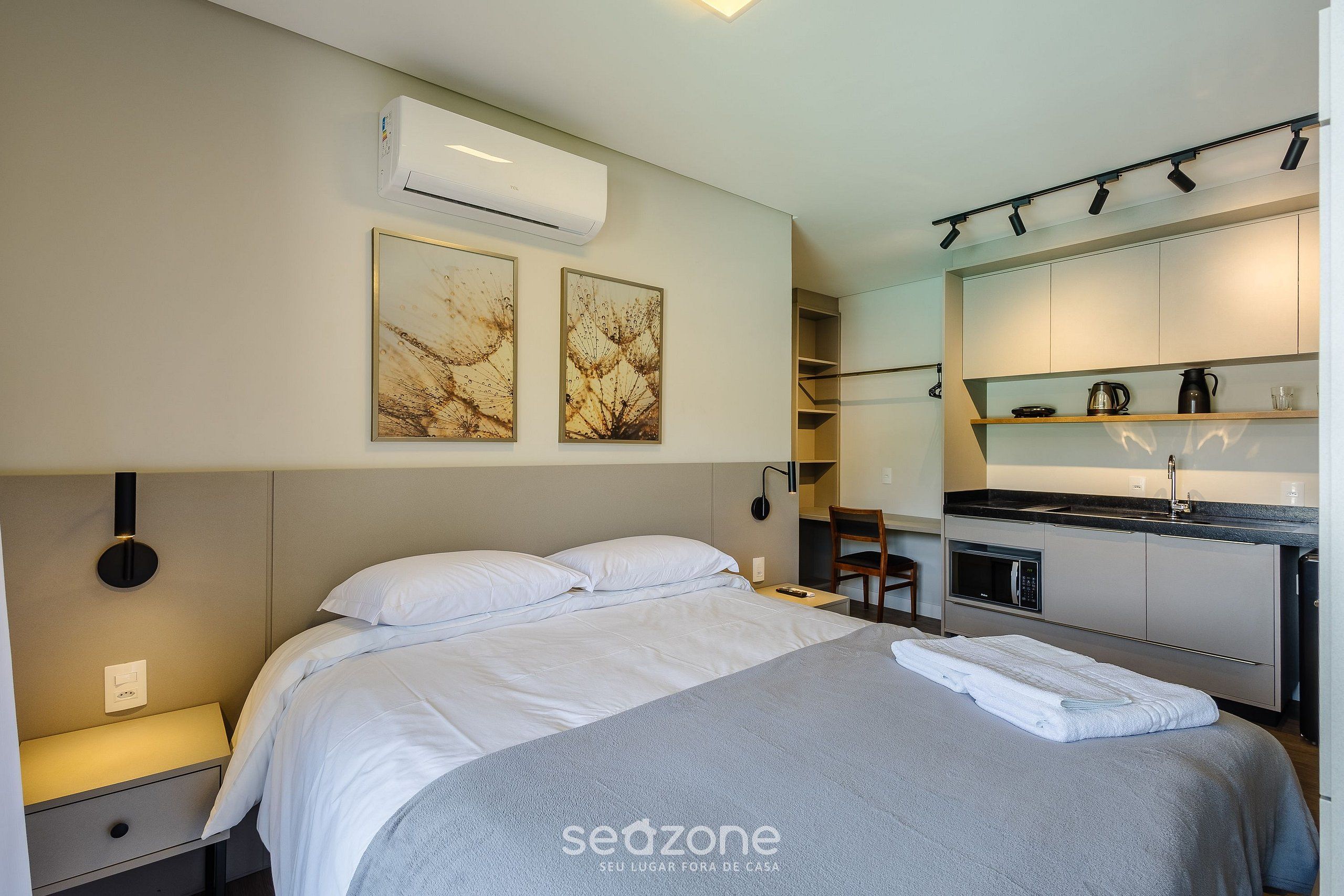 Et moderne hotelværelse med seng, sengebord, aircondition, kunstværk og et tekøkken med mikroovn og kaffemaskine.