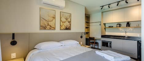 Una habitación de hotel moderna con cama, mesita de noche, unidad de aire acondicionado, obras de arte y una kitchenette con microondas y cafetera.