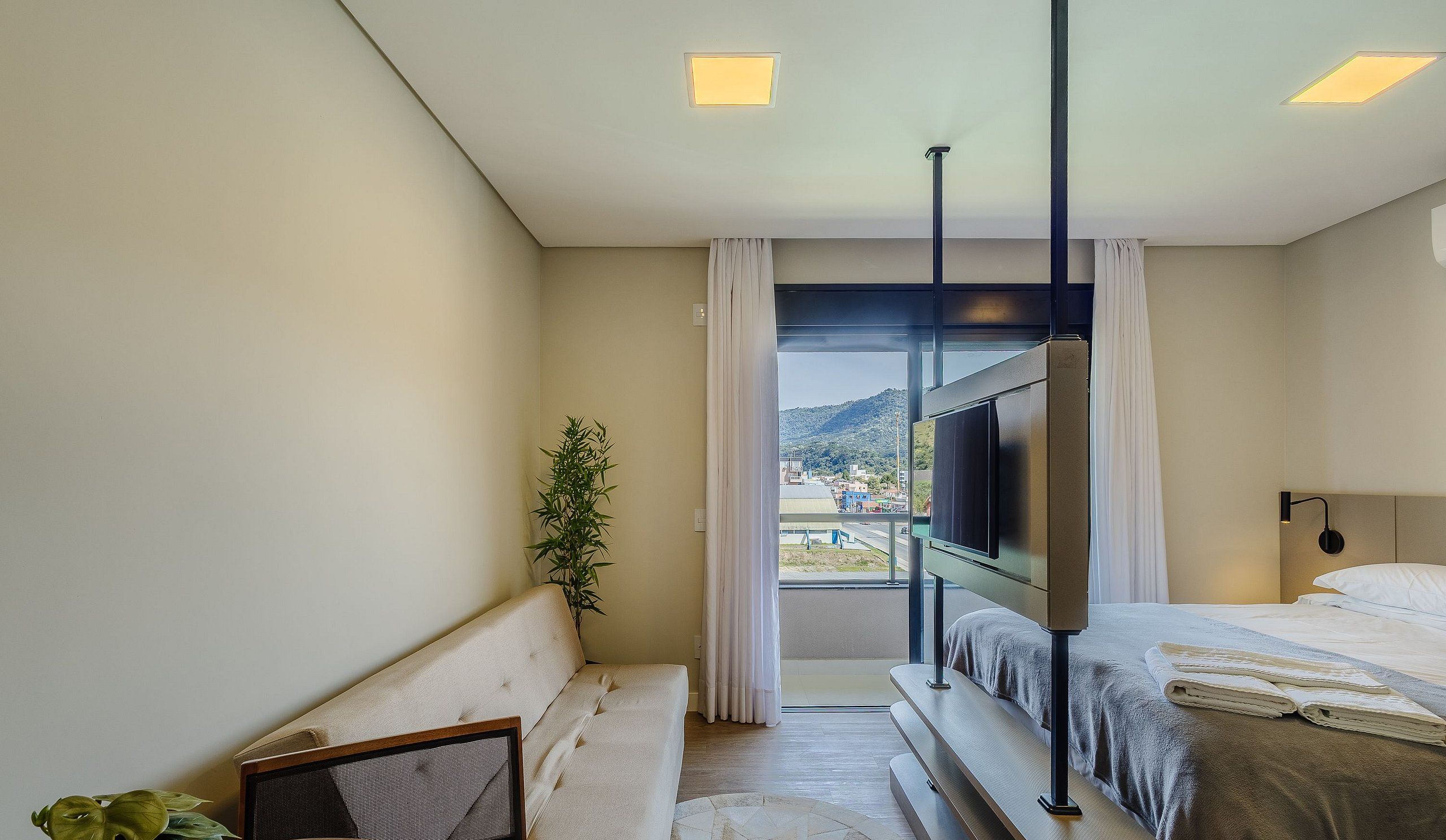 A hotel room with a bed, a TV, a plant, and a view of a cityscape.