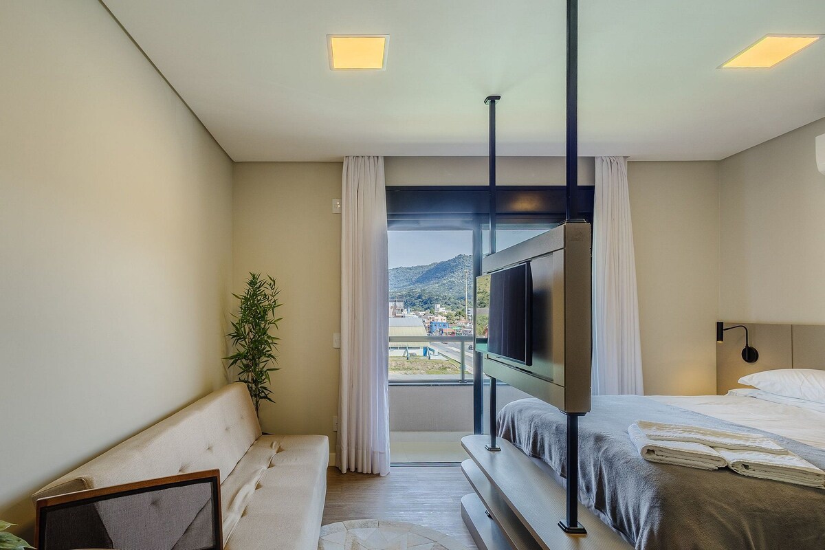 A hotel room with a bed, a TV, a plant, and a view of a cityscape.