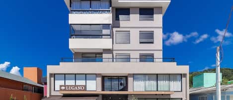 Un hotel de varios pisos con un diseño moderno, que destaca un cartel grande con la leyenda 'LUGANO' en la entrada.
