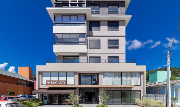 Un hotel de varios pisos con un diseño moderno, que destaca un cartel grande con la leyenda 'LUGANO' en la entrada.