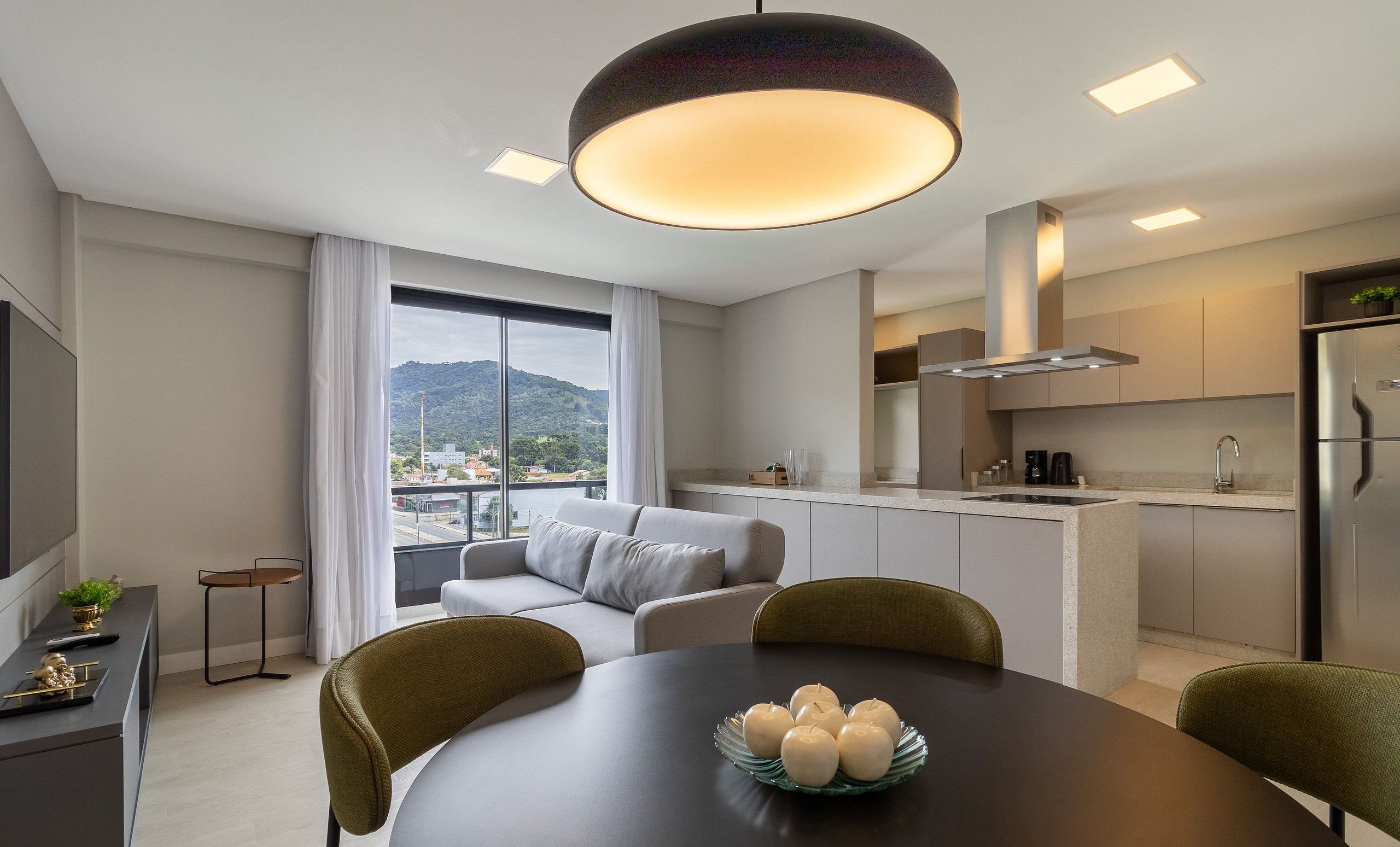 A modern kitchen with a built-in island, a dining area with a round table, and a sofa by a large window overlooking a cityscape.