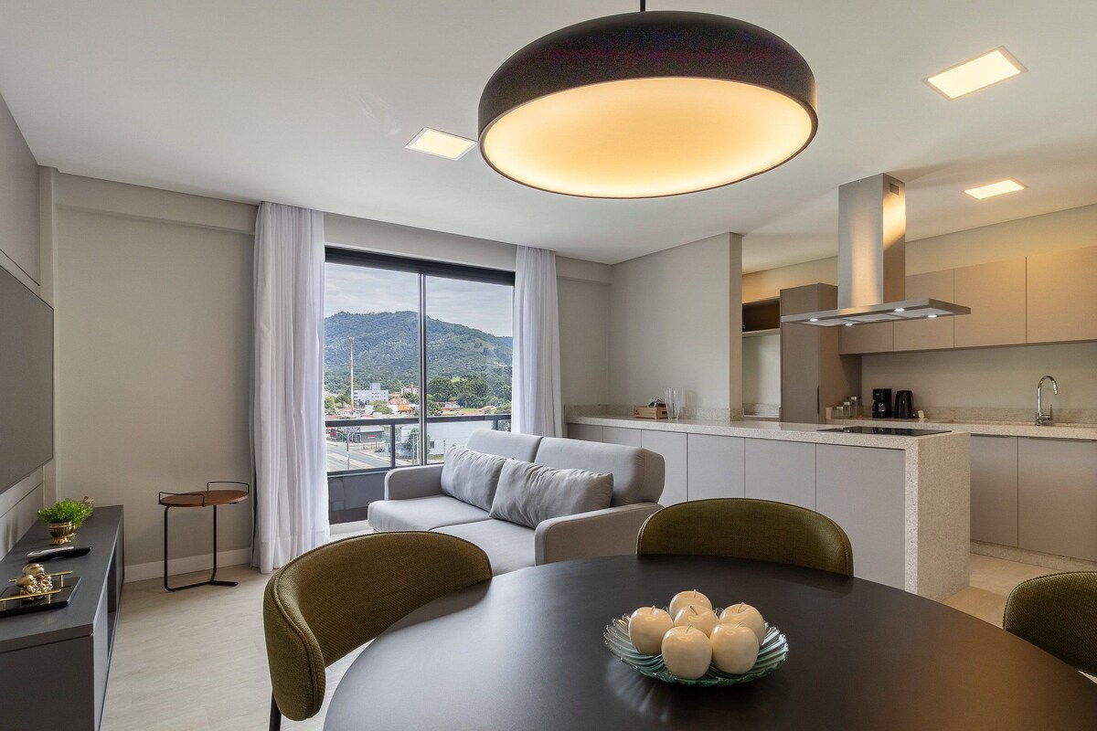 A modern kitchen with a built-in island, a dining area with a round table, and a sofa by a large window overlooking a cityscape.