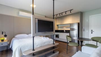 Quarto de hotel moderno e compacto com cama, televisão presa na parede, pequena cozinha e área de estar com mesa e cadeiras.