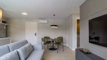 A modern living room with a grey sofa, a dining table with four chairs, a pendant light, a TV on a stand, and a kitchen area in the background.