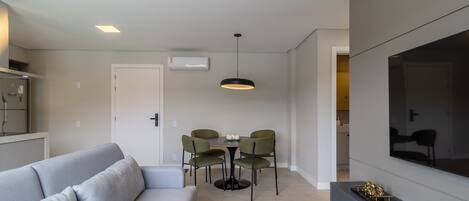 Una sala moderna con un sofá gris, una mesa de comedor con cuatro sillas, una lámpara colgante, un televisor sobre un mueble bajo y una zona de cocina al fondo.