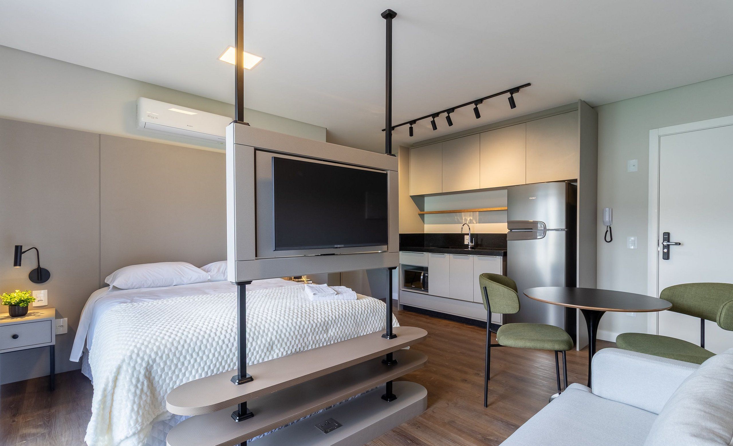 A modern, compact hotel room with a bed, a TV mounted on a wall, a small kitchenette, and a seating area with a table and chairs.