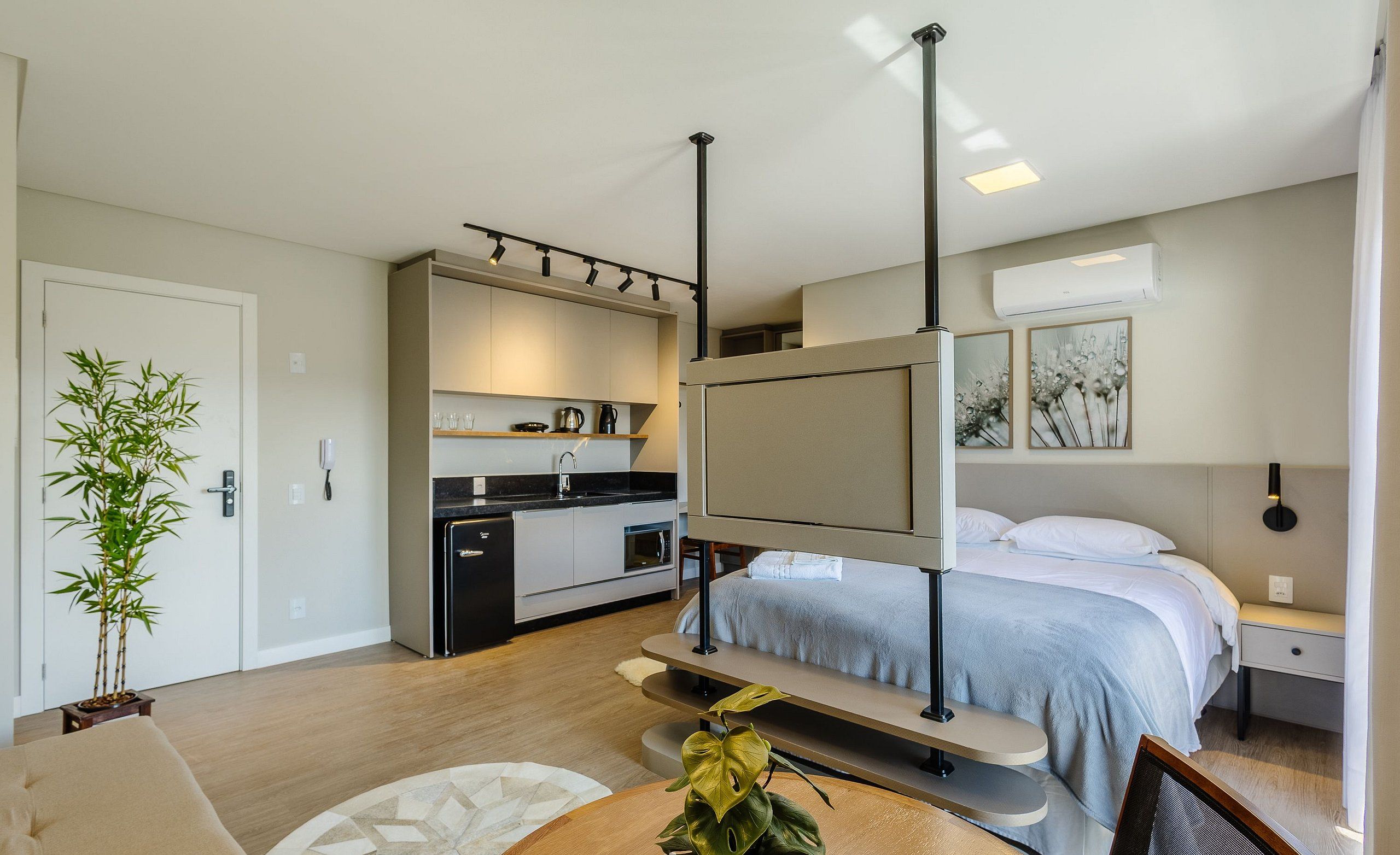 Habitación de hotel moderna con cama, mesita de noche y un pequeño escritorio con silla. Se observa una zona de cocina con pileta y alacenas. Una planta en maceta está visible en la esquina.