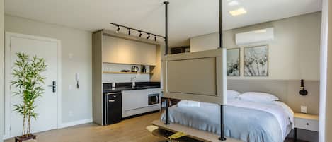 Habitación de hotel moderna con cama, mesita de noche y un pequeño escritorio con silla. Se observa una zona de cocina con pileta y alacenas. Una planta en maceta está visible en la esquina.