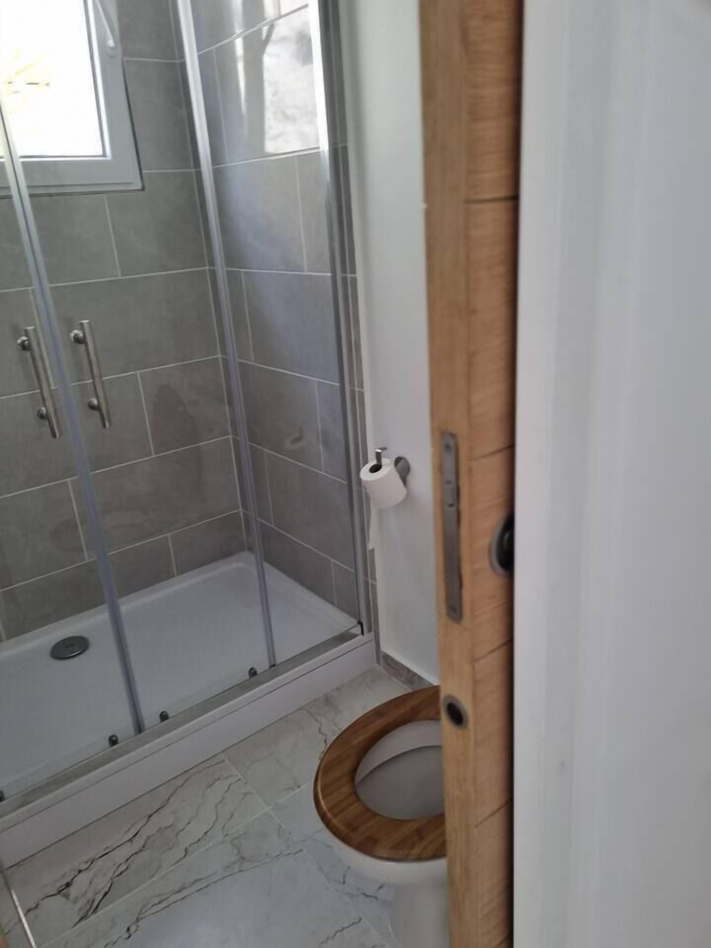 Een badkamer met een douche met glazen wand, een toilet met houten zitje en een witte zeepdispenser.