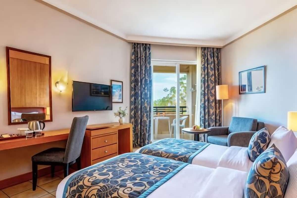 A hotel room with a desk, two beds, a TV, a balcony with curtains, and a small table with a lamp.