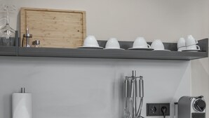 A kitchen with a wooden cabinet, a shelf with white cups and glasses, a paper towel holder, and a coffee machine. - City view apartment 10 Raspberry (Lüneburg)