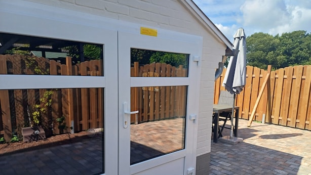 Un bâtiment blanc avec une clôture en bois, une terrasse en briques et une porte dotée d’une fenêtre.