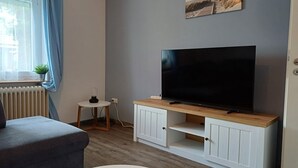 Un salon avec un téléviseur à écran plat posé sur un meuble télé en bois, un canapé gris, une petite table basse ronde et une fenêtre avec des rideaux.